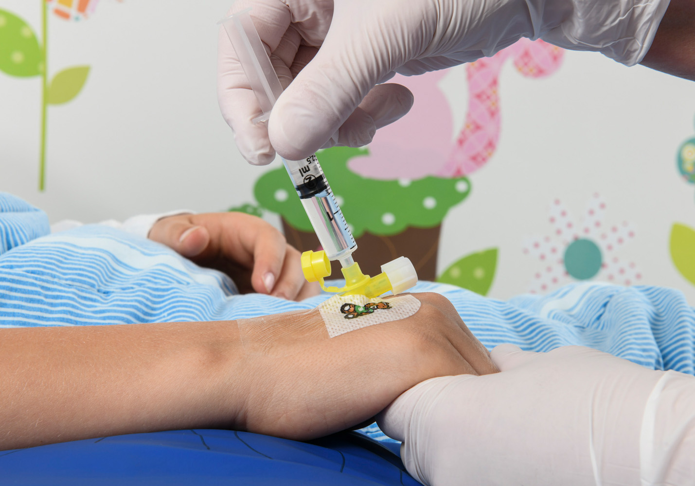 A child’s hand, with something yellow attached to the back of the hand. Somebody is injecting a transparent liquid into the yellow thing.