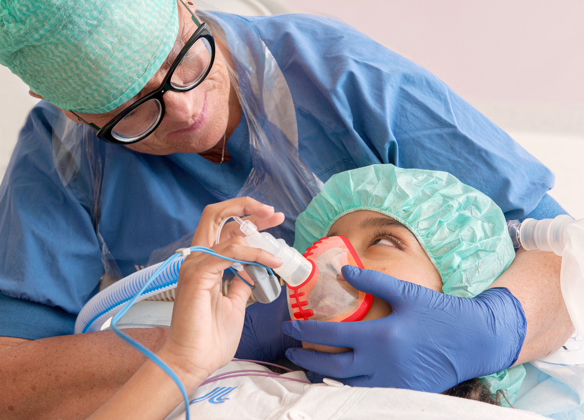 Nurse sits behind a child and holds an oxygen mask in front of the child's nose and mouth. The child and the nurse are looking at each other.
