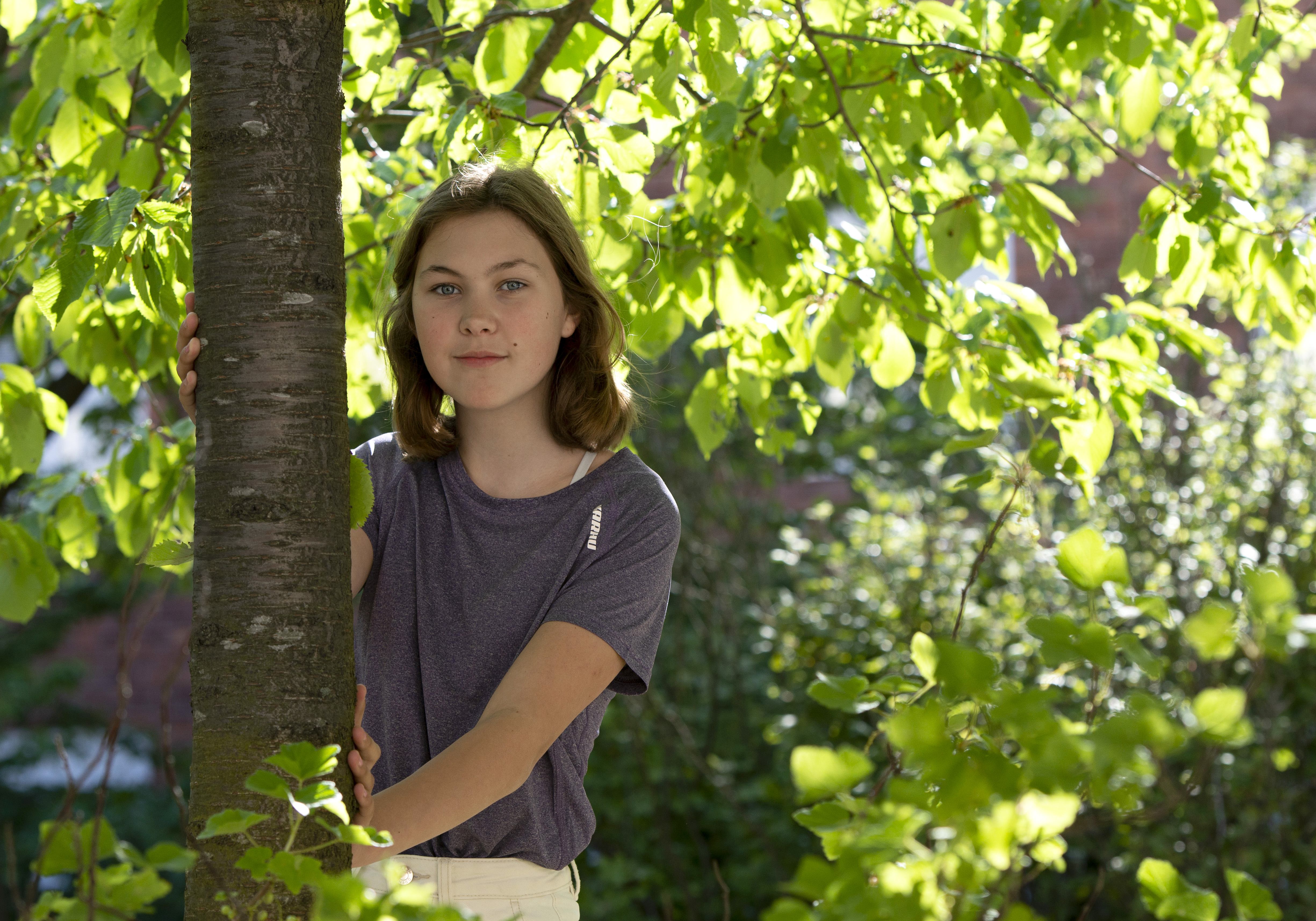 Mathilda stands leaning against a tree, with green branches and sunshine in the background.