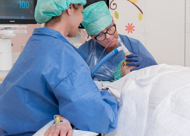 A patient on an operating table, breathing in an oxygen mask. A relative sitting beside the operating table.