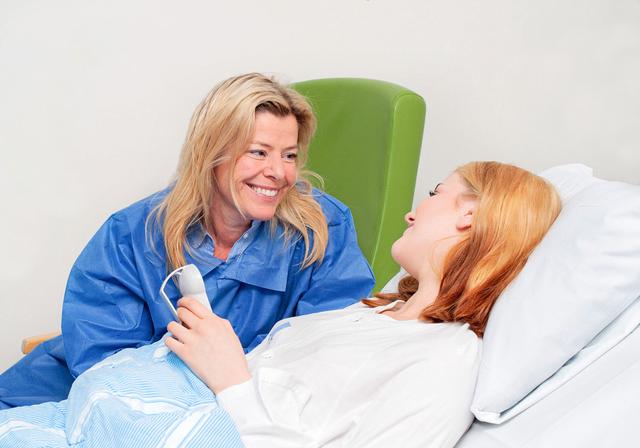 A teenage girl lying in a hospital bed. She is looking at her mother who is sitting beside the bed.